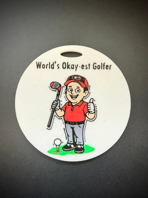 A round golf bag tag reading "World's Okay-est Golfer" with an illustrated cartoon golfer