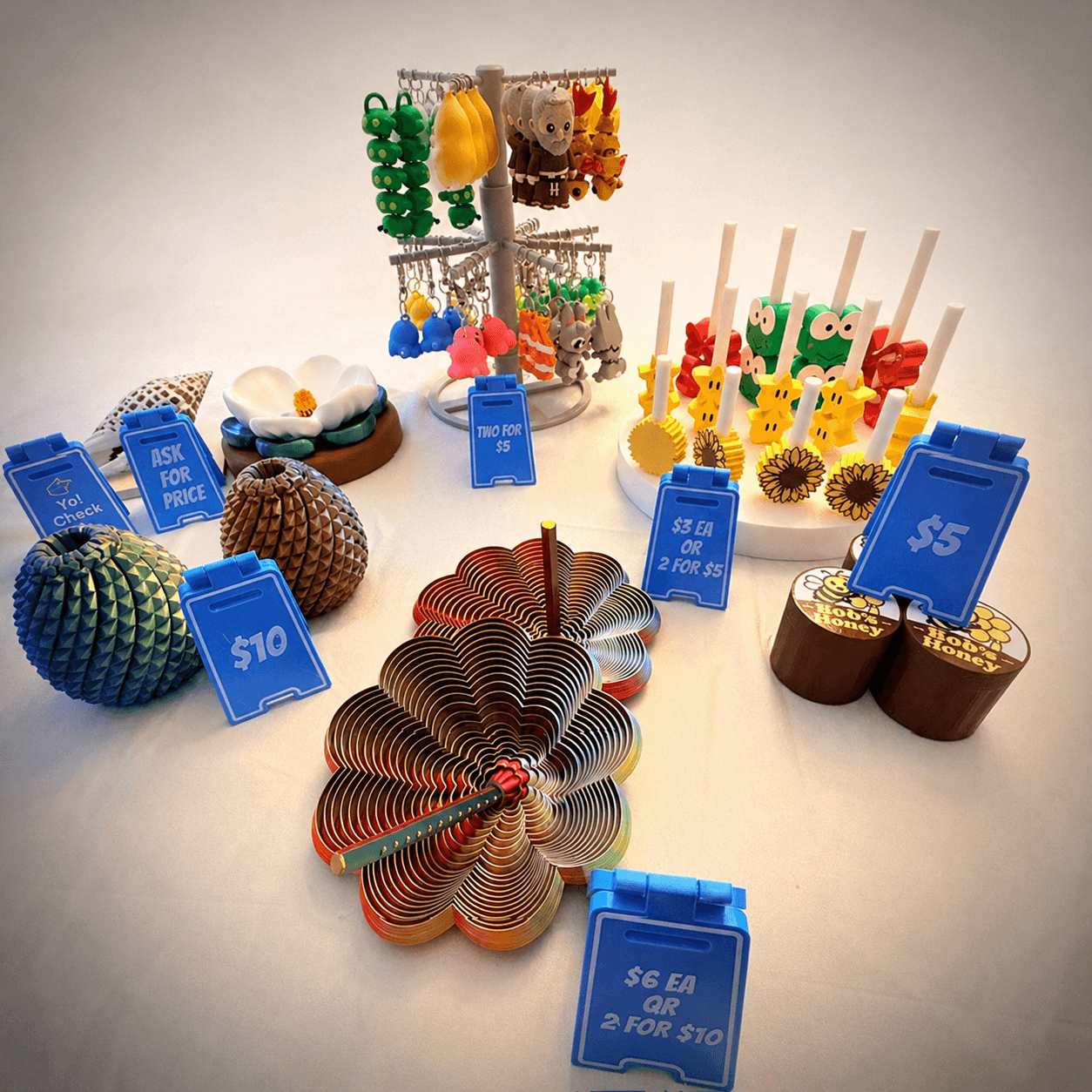 A staged demo table with 3D-printed items, keychains, and custom price signs, illustrating a market display concept