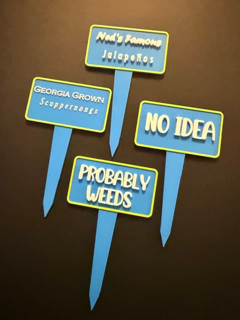 Four custom garden stake signs, including "Ned's Famous Jalapeños," "Georgia Grown Scuppernongs," "No Idea," and "Probably Weeds"
